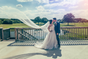 Bride and groom stood on the balcony at Sandburn Hall in York, with the vail blowing in the wind. Wedding Photography by Charlotte Atkinson